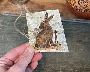 A handmade gift tag. It is a piece of real birch bark with a woodland rabbit wood burned onto the bark. A jute cord is tied in the corner.